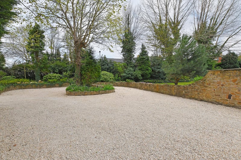 From High Street, a wide opening gives access to a gravelled driveway with a turning circle around a raised stone planter with mature trees and planted shrubs. The driveway provides parking for several vehicles with exterior lighting and mature tree/shrub borders.